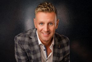 Michael English, the Irish singer, smiles warmly at the camera. He’s wearing a grey checkered blazer and white shirt, with short blonde hair and bright blue eyes, set against a dark background.