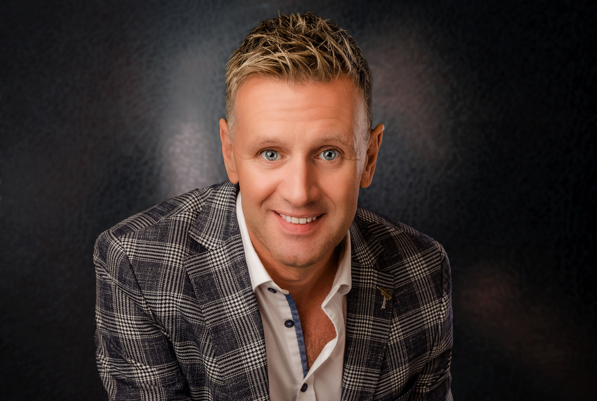 Michael English, the Irish singer, smiles warmly at the camera. He’s wearing a grey checkered blazer and white shirt, with short blonde hair and bright blue eyes, set against a dark background.