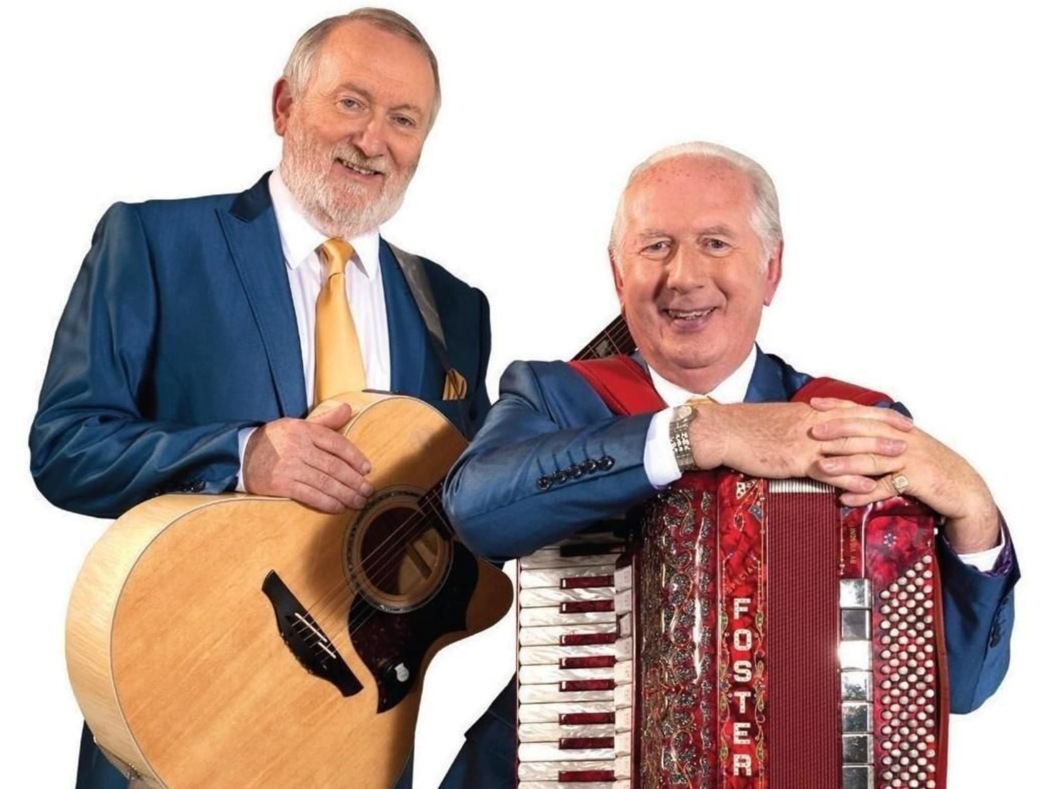 Foster and Allen, the Irish musical duo, are shown smiling in this image. One, holding a guitar, wears a blue suit and gold tie, while the other leans on a red accordion labeled 'Foster,' also dressed in a matching blue suit and gold tie. Both men appear joyful and charismatic.