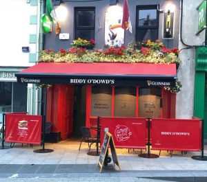 Front view of Biddy O'Dowd's pub with a red canopy, floral display, and signs for live music and food offerings.